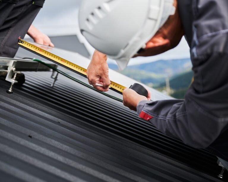 Back view of competent technician making measurements for exact calculations. Adjusting PV solar panel on a roof. Engineer in helmet making calculations of solar cell with a measuring tape.