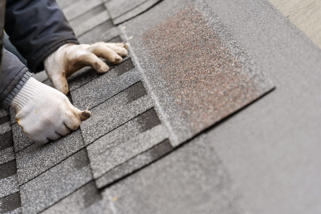 Close up and real photo of skilled workman in special protective work wear installing asphalt or bitumen tile on top of the roof under construction house