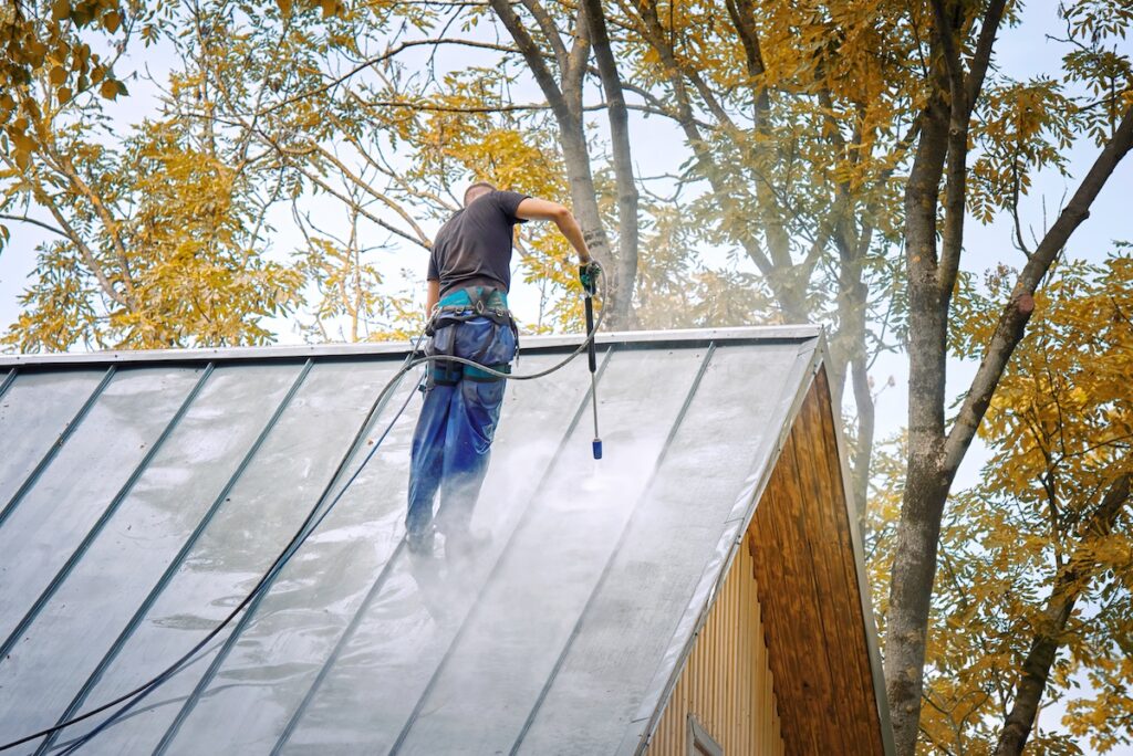 Man with pressure washer gun washing roof of private house. Cleaner with high pressure water sprayer washes steel roof from dirt. Handyman cleaning roof with water pressure cleaner.