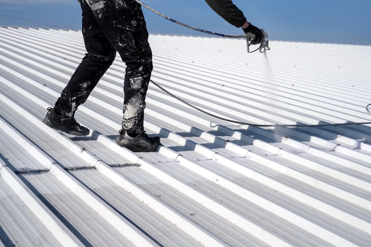 Metal roof painting. Technician performing painting of roof in white color. Work at height. Roof repairing service. Rooftop maintenance.