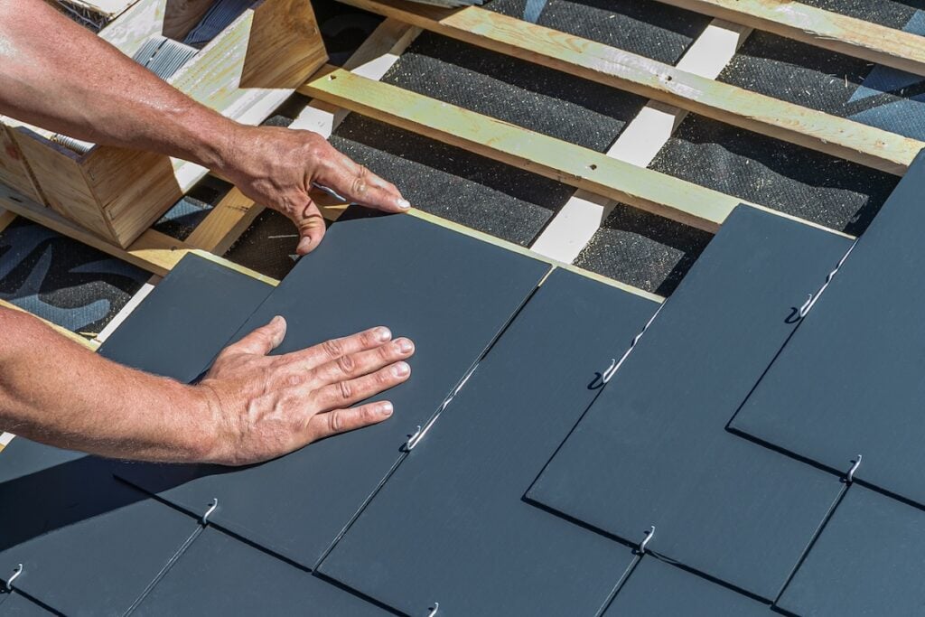 Worker installs gray slate roof tiles with metal hook fasteners