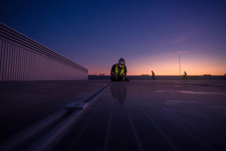 Technician inspecting flat commercial roof at dusk with crew nearby