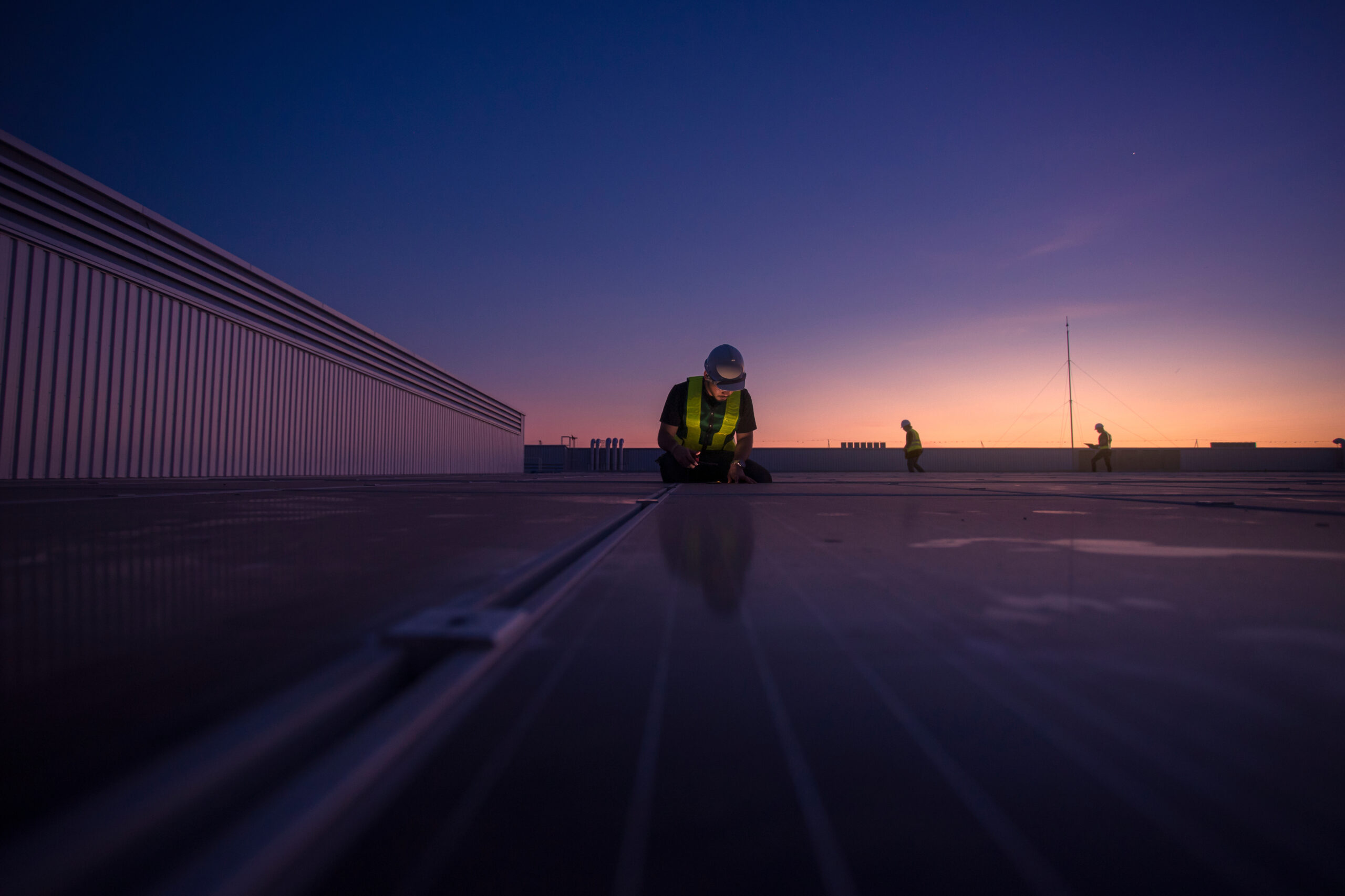 Technician inspecting flat commercial roof at dusk with crew nearby