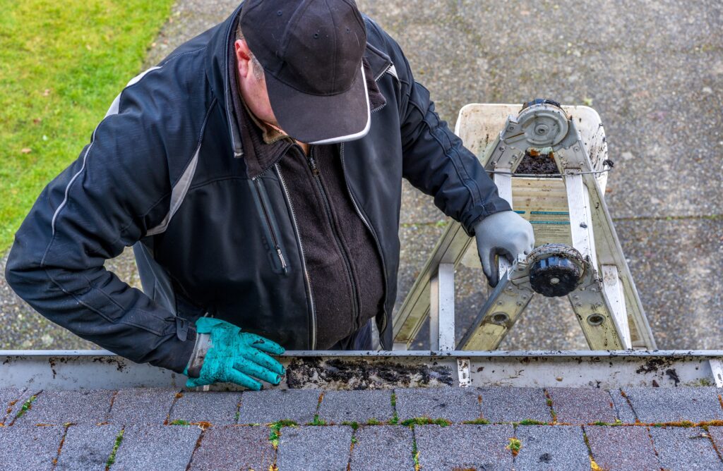 Worker removing debris from clogged gutter during roof maintenance visit