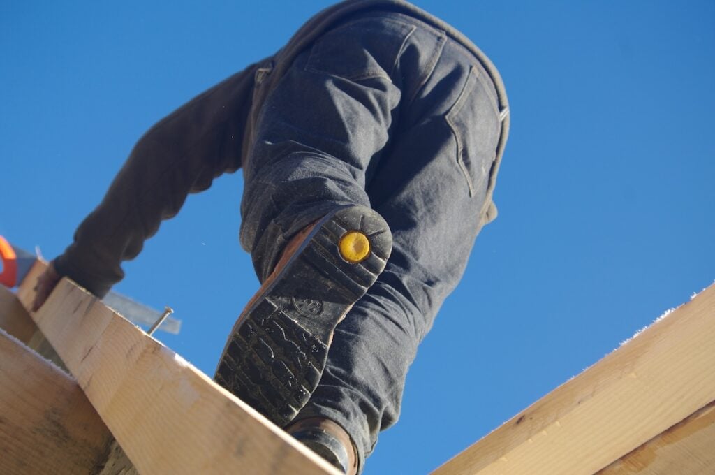 Roofer climbing wood rafters in slip-resistant work boots against blue sky