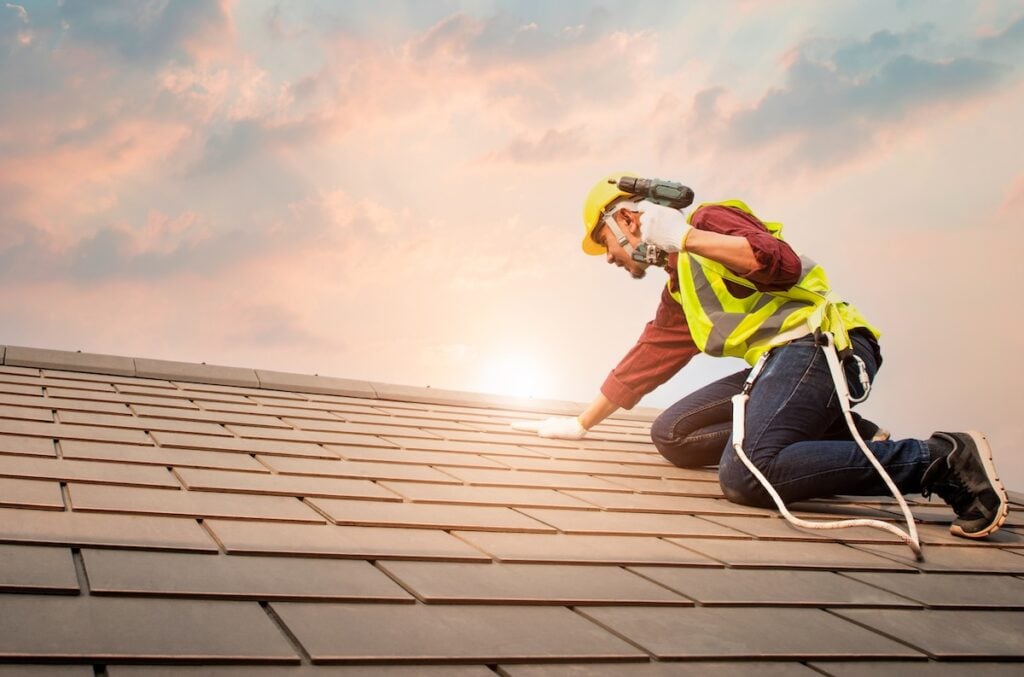 Roofer kneeling on tile roof wearing safety harness and grip-sole boots