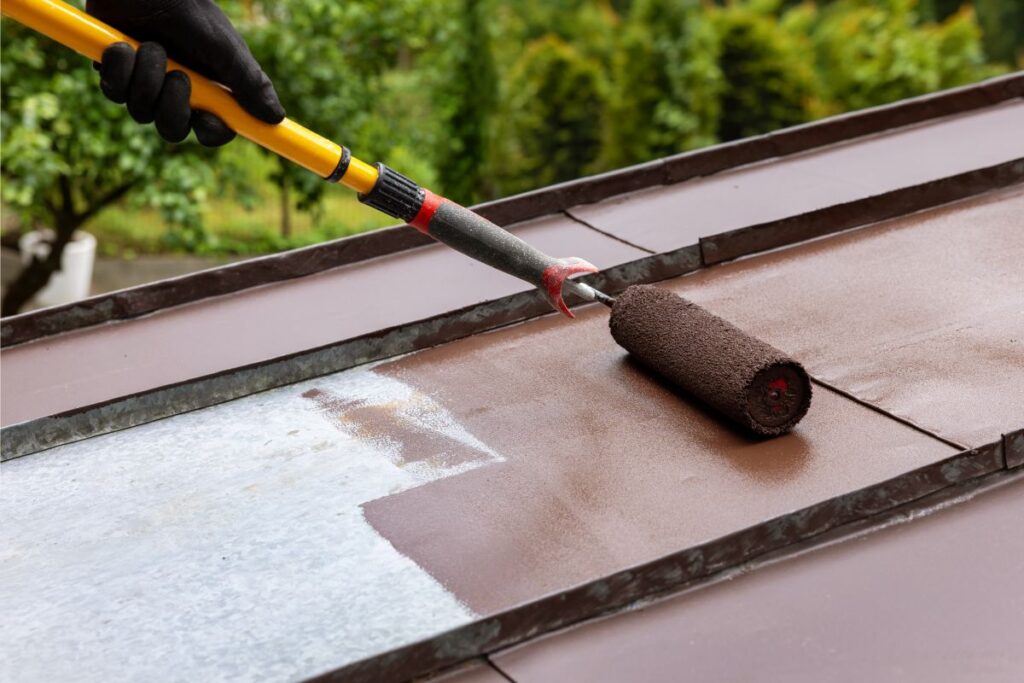 man painting tin metal roof with roller and brown paint. house roofing restoration
